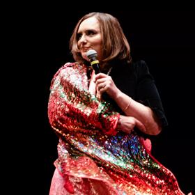 Deborah Frances-White, host of the award winning podcast 'The Guilty Feminist', on stage holding a microphone wearing  a sparkly shall.