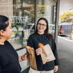 Two people enjoy hot cross buns outside of a bakery.