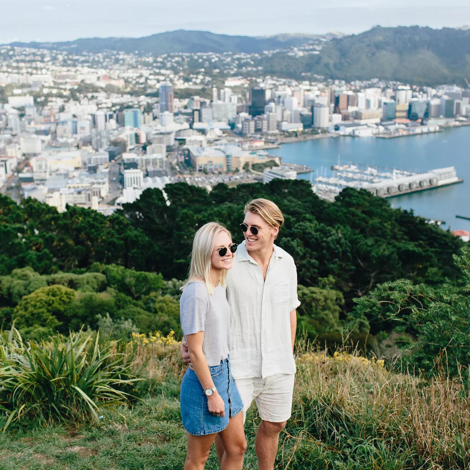 2 people stand close together atop Mount Victoria, and the city of Wellington can be seen behind.