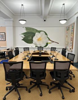 A boardroom at the New Zealand Parliament with desks, swivel chairs and a mural of a camellia flower on the back wall.