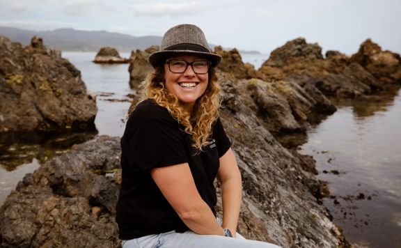 Founder of Octacle, Kat Greagor, sits on a rock at the beach. She wears a grey fedora, a black tee shirt and light blue jeans, smiling.