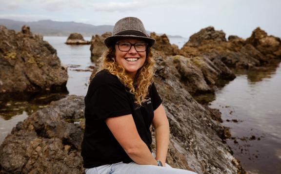 Founder of Octacle, Kat Greagor, sits on a rock at the beach. She wears a grey fedora, a black tee shirt and light blue jeans, smiling. 