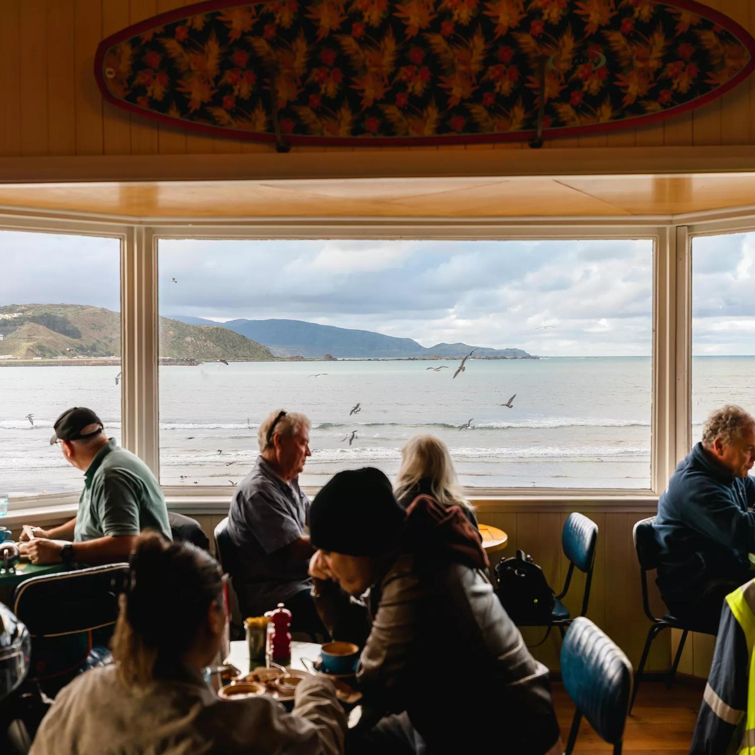 The interior of the iconic Maranui Cafe in Lyall Bay, Wellington. The bay, overcast sky and flying gulls are seen through the three-pane bay window. There are four tables with two patrons sitting at each. 