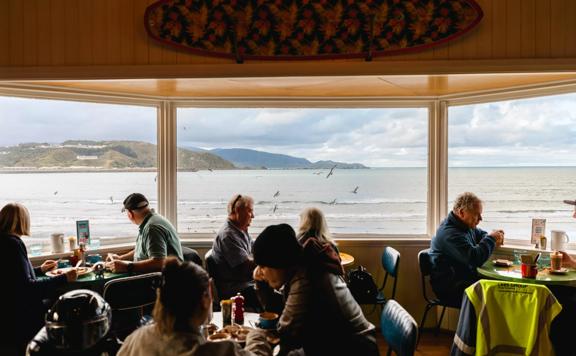 The interior of the iconic Maranui Cafe in Lyall Bay, Wellington. The bay, overcast sky and flying gulls are seen through the three-pane bay window. There are four tables with two patrons sitting at each. 