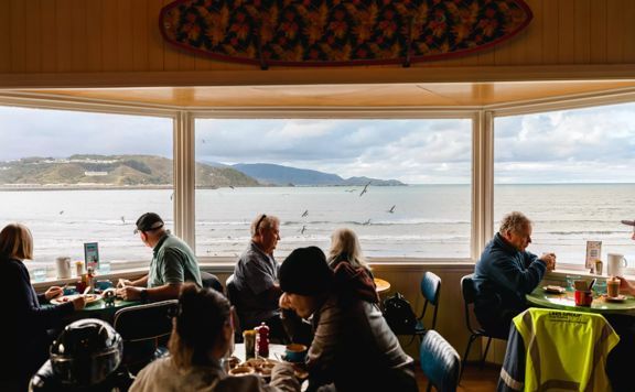 The interior of the iconic Maranui Cafe in Lyall Bay, Wellington. The bay, overcast sky and flying gulls are seen through the three-pane bay window. There are four tables with two patrons sitting at each.