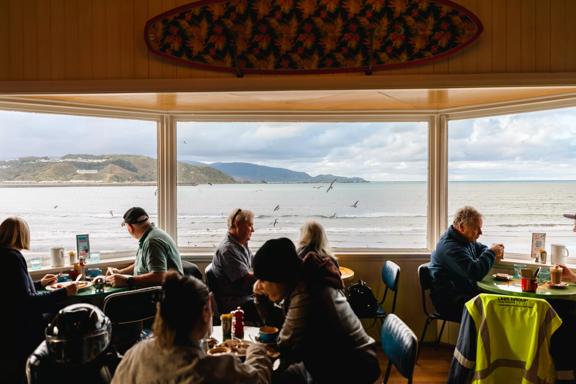 The interior of the iconic Maranui Cafe in Lyall Bay, Wellington. The bay, overcast sky and flying gulls are seen through the three-pane bay window. There are four tables with two patrons sitting at each. 