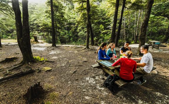 five people sat on a bench at Butterfly creek enjoying a picnic amongst the shady trees.