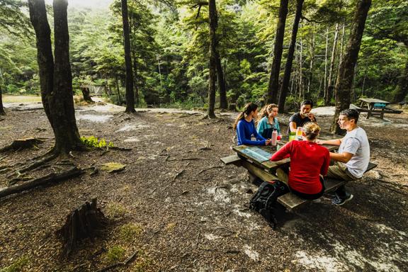 five people sat on a bench at Butterfly creek enjoying a picnic amongst the shady trees.