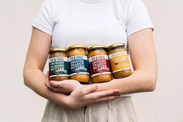 A person wearing white holds a collection of Fix & Fogg peanut butter jars in their arms