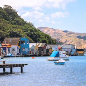 Colourful boatsheds line the edge of a blue body of water with rolling hills in the background.