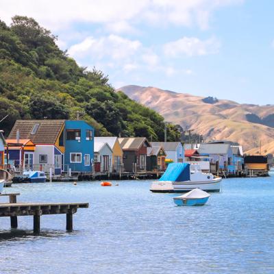 Colourful boatsheds line the edge of a blue body of water with rolling hills in the background.