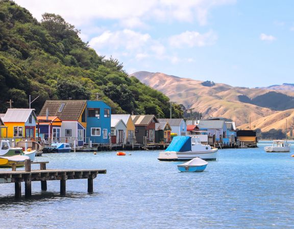 Colourful boatsheds line the edge of a blue body of water with rolling hills in the background.