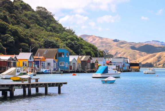 Colourful boatsheds line the edge of a blue body of water with rolling hills in the background.