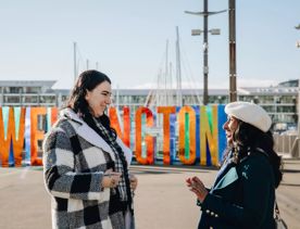 Two smiling friends chatting while standing in front of the Well_ngton Sign at the city's harbourfront.