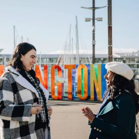 Two smiling friends chatting while standing in front of the Well_ngton Sign at the city's harbourfront.