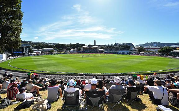 An ongoing cricket match at Cello Basin Reserve under a sunny blue sky. Fans sit on the grass and lawn chairs to watch.