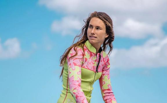 A person sits on a paddleboard, holding a paddle. She wears a colourful wet suit.