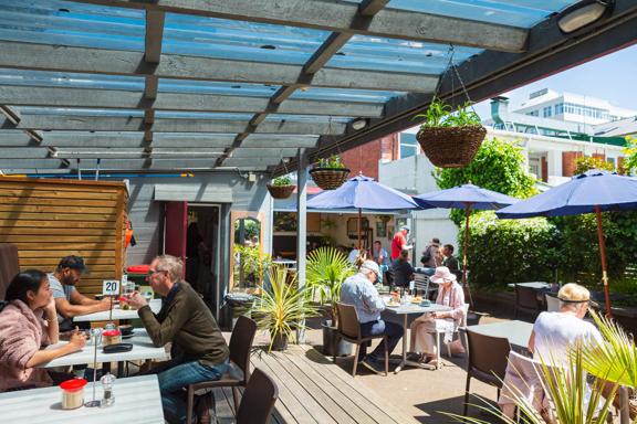 People sit at tables on the sun-soaked deck at Hive. Blue sun umbrellas provide shade.