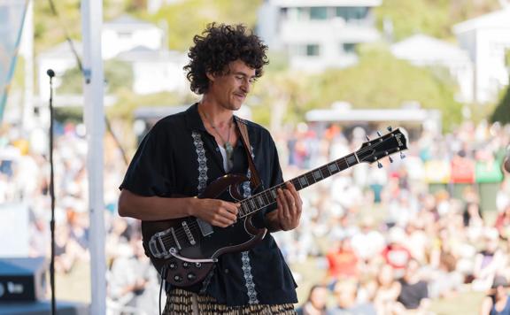 Person playing an electric guitar on a stage with a crowd in the background.