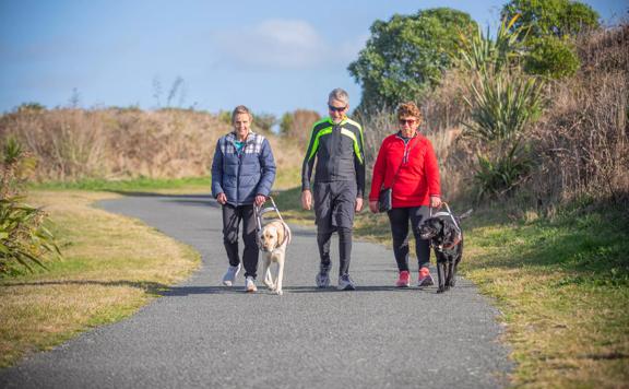 Three people walk along the sealed Te Ara o Whareroa path in Queen Elizabeth Park. Two of the people are being assisted by Guide Dogs.
