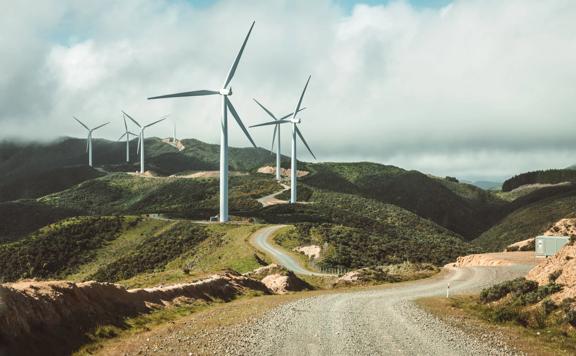 Eight large wind turbines set on rolling hills with a gravel road winding around them.