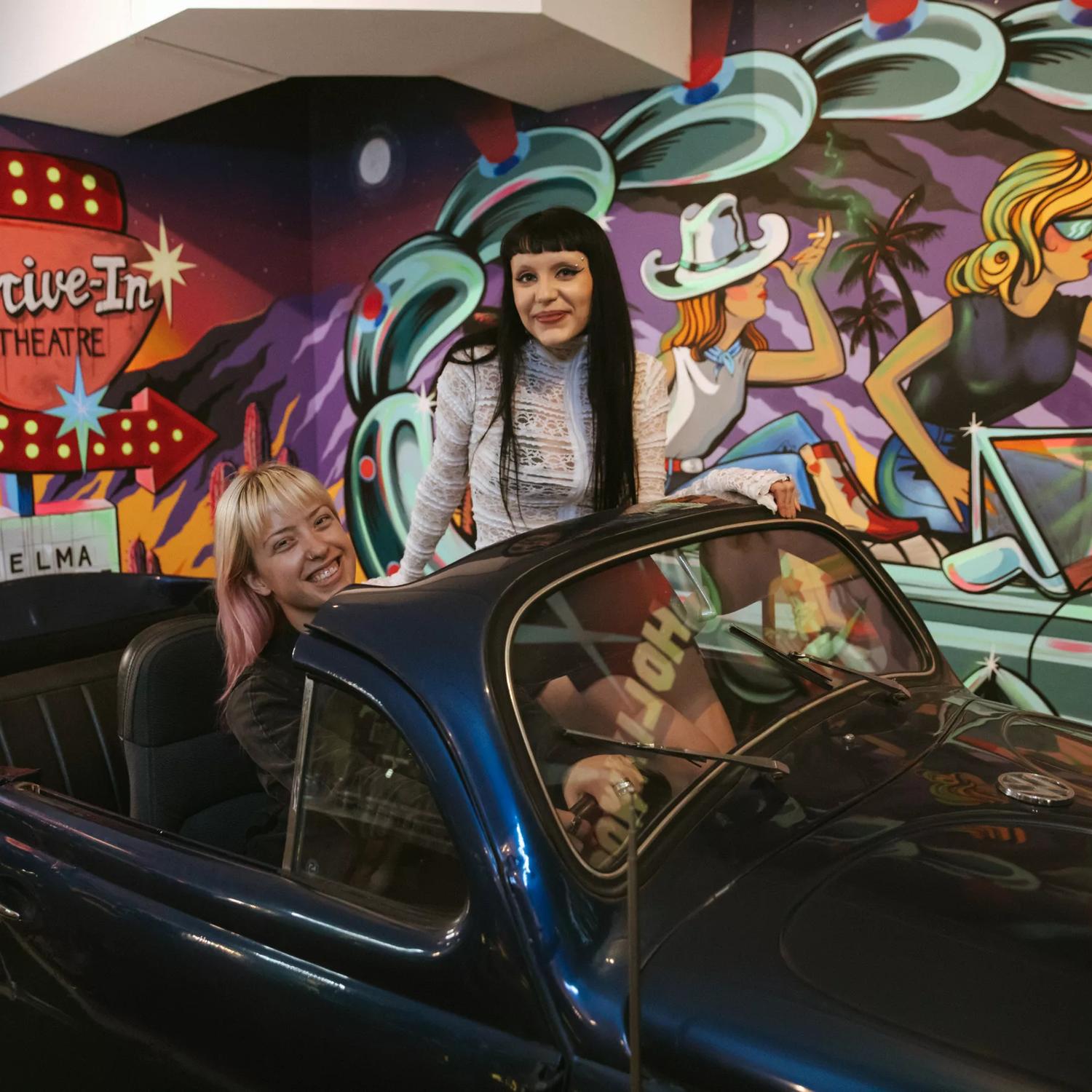 Inside Bizarre Bazaar, a thrift store in Te Aro, Wellington. Two people are sitting in an old convertible car with a colourful Americana mural on the wall behind them. 