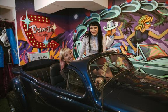 Inside Bizarre Bazaar, a thrift store in Te Aro, Wellington. Two people are sitting in an old convertible car with a colourful Americana mural on the wall behind them. 