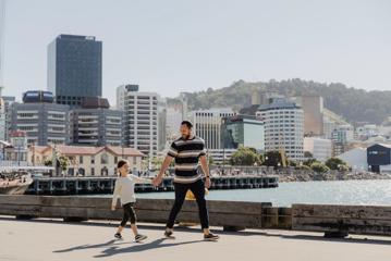 Parent and child walk along the waterfront, with the rowing club and Wellington central buildings seen behind them.