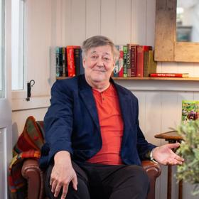 Stephen Fry sits in a leather armchair wearing an orange shirt and navy blue blazer.