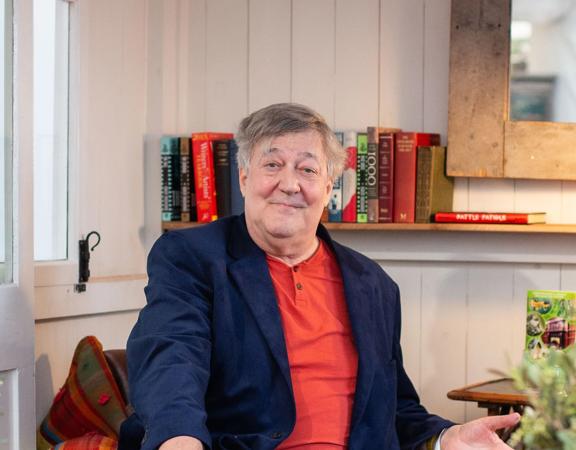 Stephen Fry sits in a leather armchair wearing an orange shirt and navy blue blazer. 