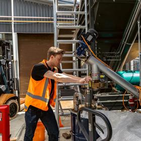 A person in an bright orange safety vest is working on the a metal machine in an industrial warehouse.