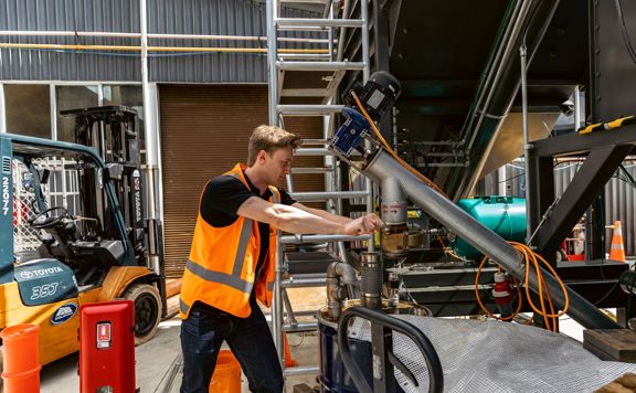 A person in an bright orange safety vest is working on the a metal machine in an industrial warehouse.