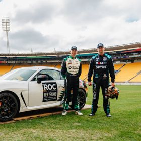 Two race drivers stand beside a white race car on the field of a large stadium.
