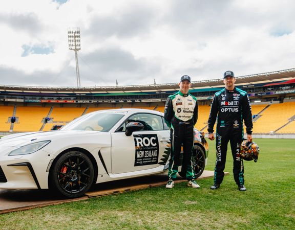 Two race drivers stand beside a white race car on the field of a large stadium.