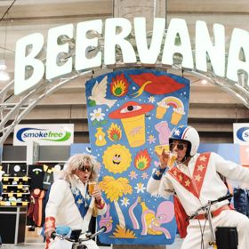 Two people wearing white leather jumpsuits with blue and red star details do silly poses on bicycles under a Beervana sign.