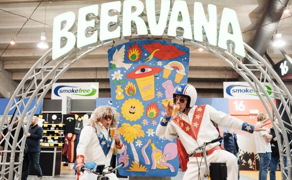 Two people wearing white leather jumpsuits with blue and red star details do silly poses on bicycles under a Beervana sign.
