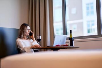 A person sitting at a table, talking on their cellphone with a glass of red wine and a laptop on the table at Sojourn Apartment Hotel in Wellington, New Zealand.