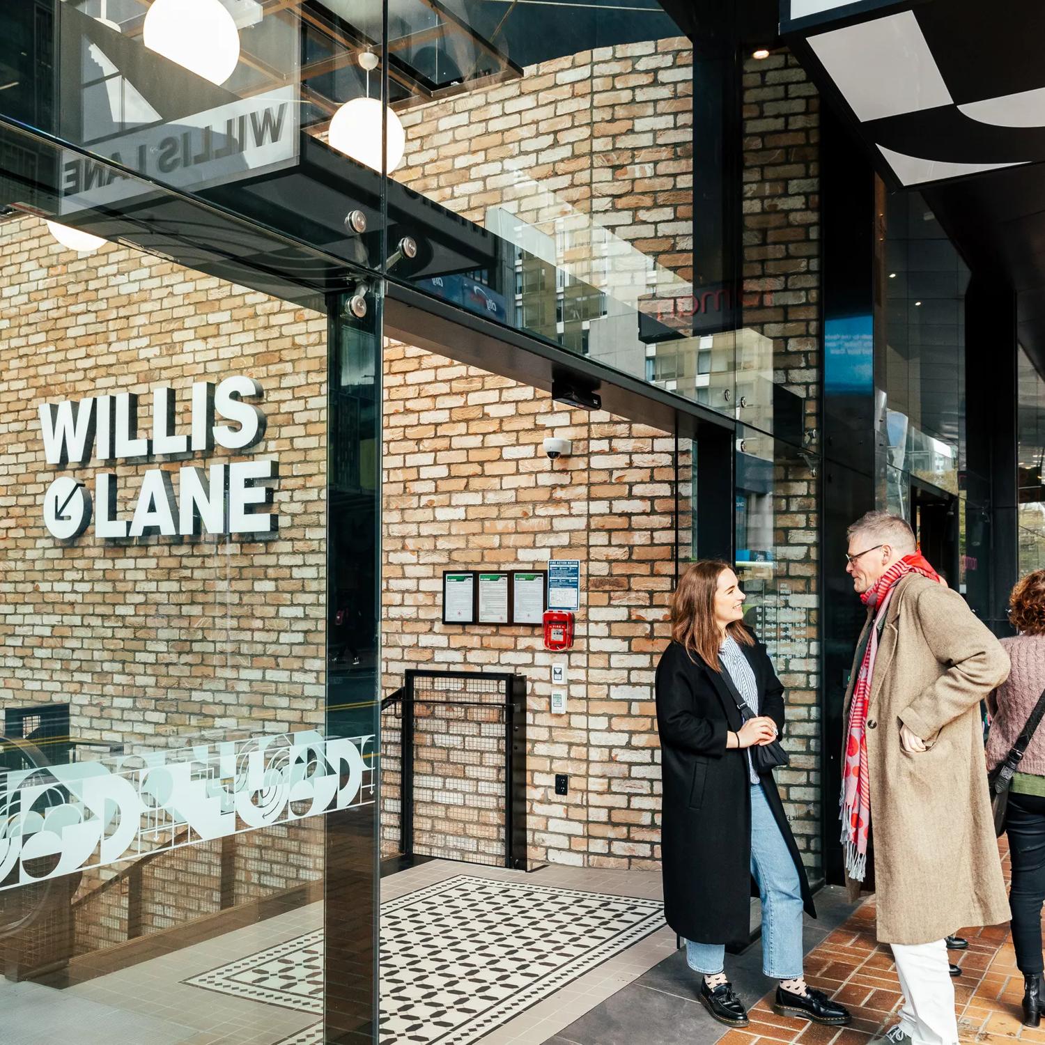 People walking by and standing outside the entry to Willis Lane, with its large glass window, sliding door, brick wall, and sign leading down to Willis Lane.