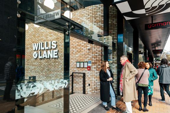 People walking by and standing outside the entry to Willis Lane, with its large glass window, sliding door, brick wall, and sign leading down to Willis Lane.