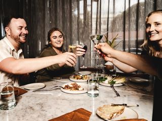 People seated around a table raise glasses in a toast during a meal, with plates of food and drinks visible in front of them.