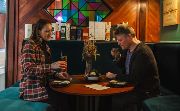 Two people sit at a circular table on green velvet seats holding cocktails.