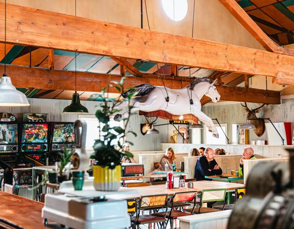 The interior of Seashor Cabaret, a diner located on The Esplanade and Sidney Street in Petone, with four vintage arcade games, two taxidermy deer heads on the wall, two large tables with eight chairs each and three wood booths with eight customers seated.