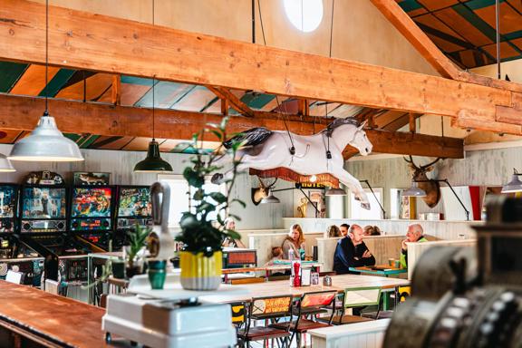 The interior of Seashor Cabaret, a diner located on The Esplanade and Sidney Street in Petone, with four vintage arcade games, two taxidermy deer heads on the wall, two large tables with eight chairs each and three wood booths with eight customers seated.