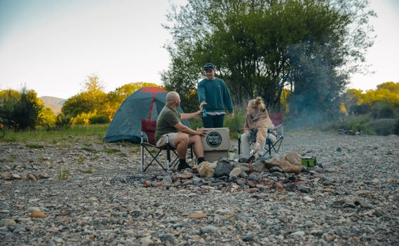 Three friends enjoy some drinks by a fire while camping at Kaitoke Regional Park.