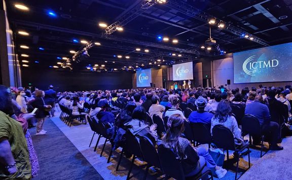 People sitting on rows of chairs in a large conference space, facing towards projector screens that have the ICTMD logo on display.
