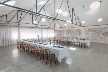 The interior of Whisky and Wood, an event venue in Wellington Central, setup with two long banquet tables, with white table clothes and twenty wooden chairs at each, exposed brick walls and circular lights suspended from the ceiling.