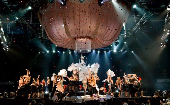 In the 2006 World of WearableArt show, people are dancing on stage with large feathers as confetti rained down. A large chandelier made of pink material hangs above them.