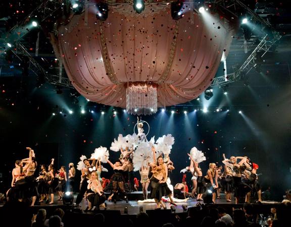 In the 2006 World of WearableArt show, people are dancing on stage with large feathers as confetti rained down. A large chandelier made of pink material hangs above them.