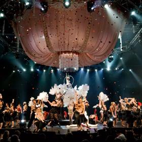 In the 2006 World of WearableArt show, people are dancing on stage with large feathers as confetti rained down. A large chandelier made of pink material hangs above them.