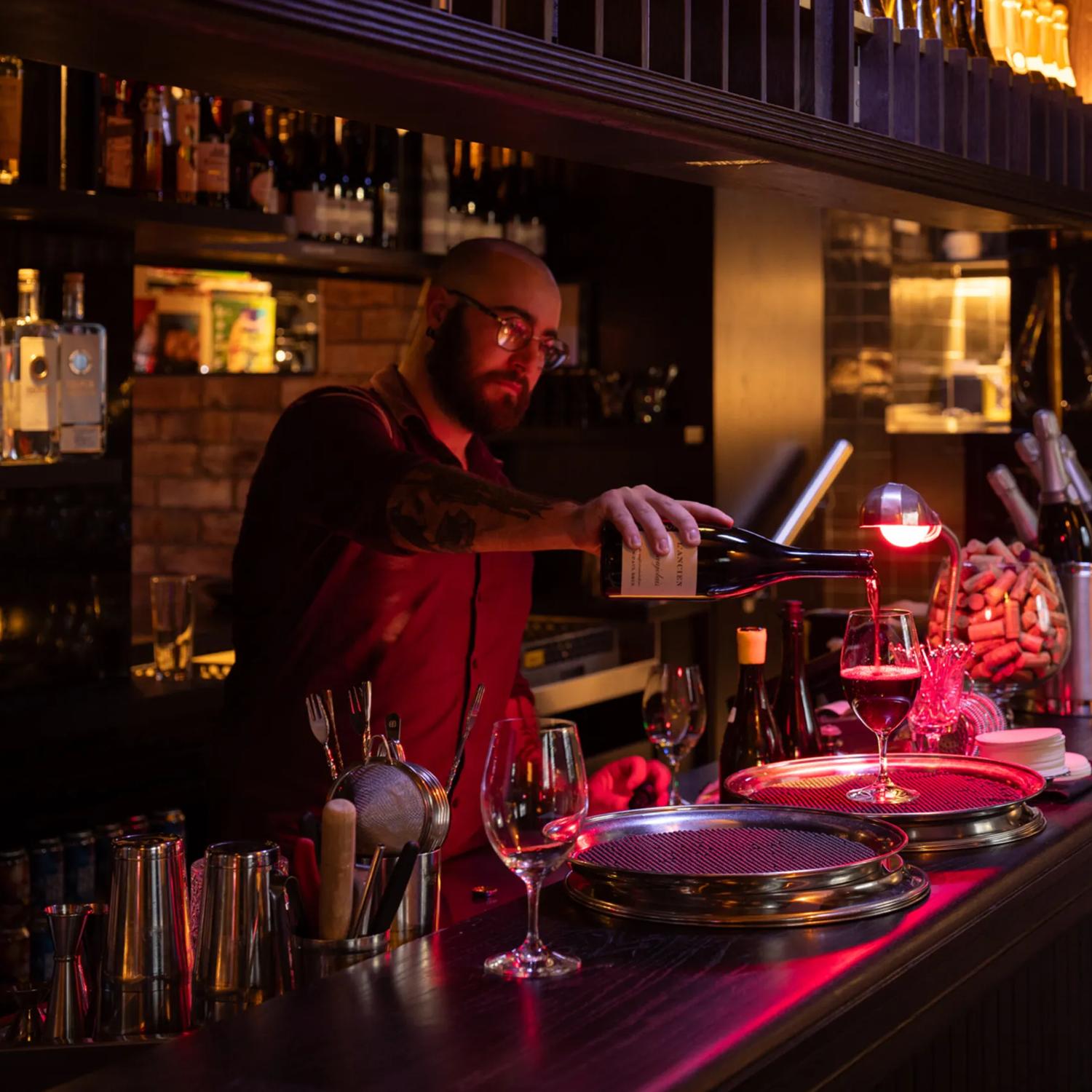 A bartender pours a glass of red wine in a dark cozy wine bar.